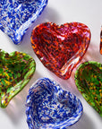 Six heart-shaped glass bowls in assorted colors and patterns, including blue-and-white speckled, red-and-orange marbled, and green-and-yellow mottled designs, arranged on a white background.