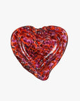 A heart-shaped glass dish with a textured, bubbled pattern in vibrant red, orange, and purple tones, set against a plain light gray background.