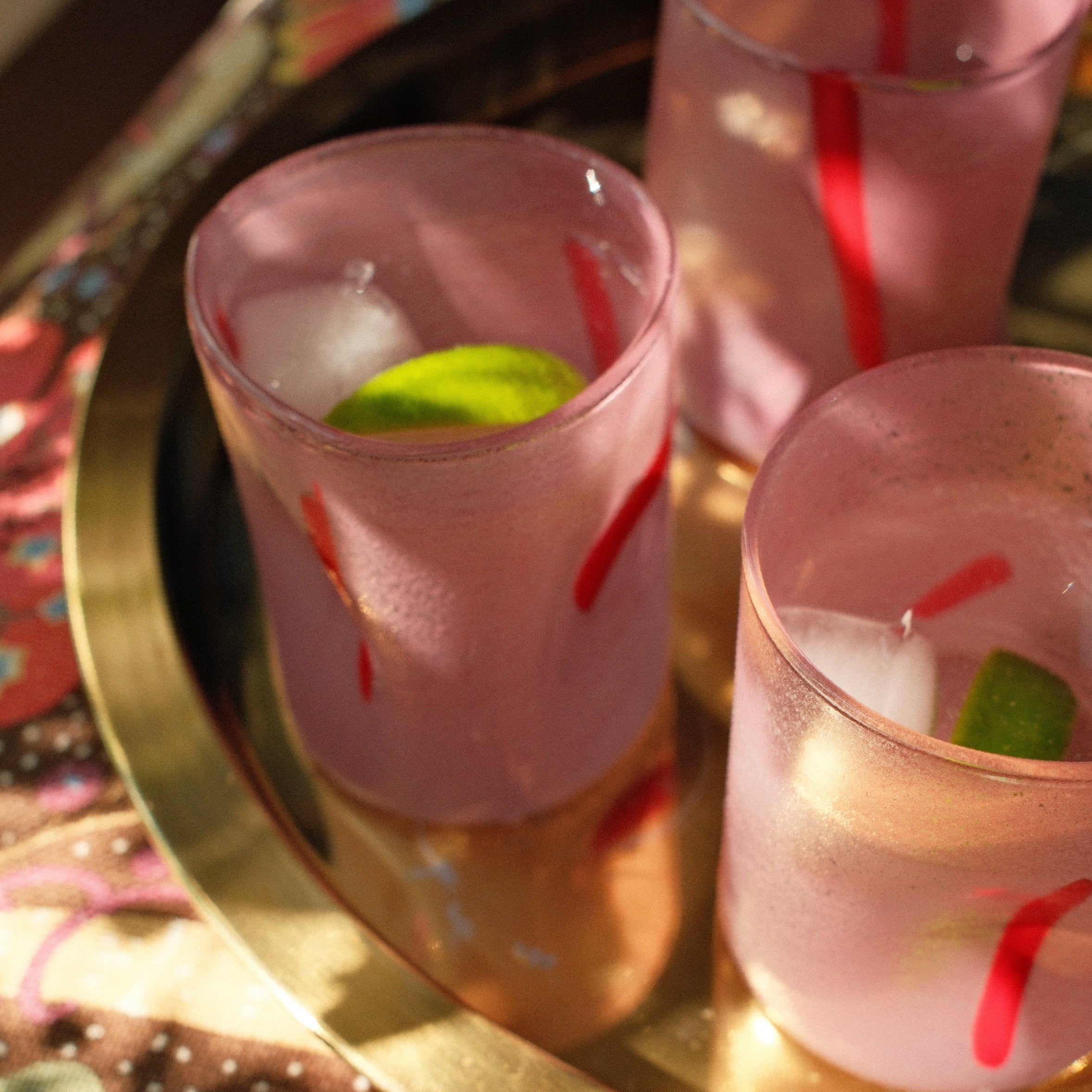 Three frosted pink glasses with red stripes and lime wedges, filled with a pale pink drink and ice, arranged on a round gold tray over a floral tablecloth.