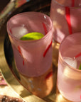 Three frosted pink glasses with red stripes and lime wedges, filled with a pale pink drink and ice, arranged on a round gold tray over a floral tablecloth.