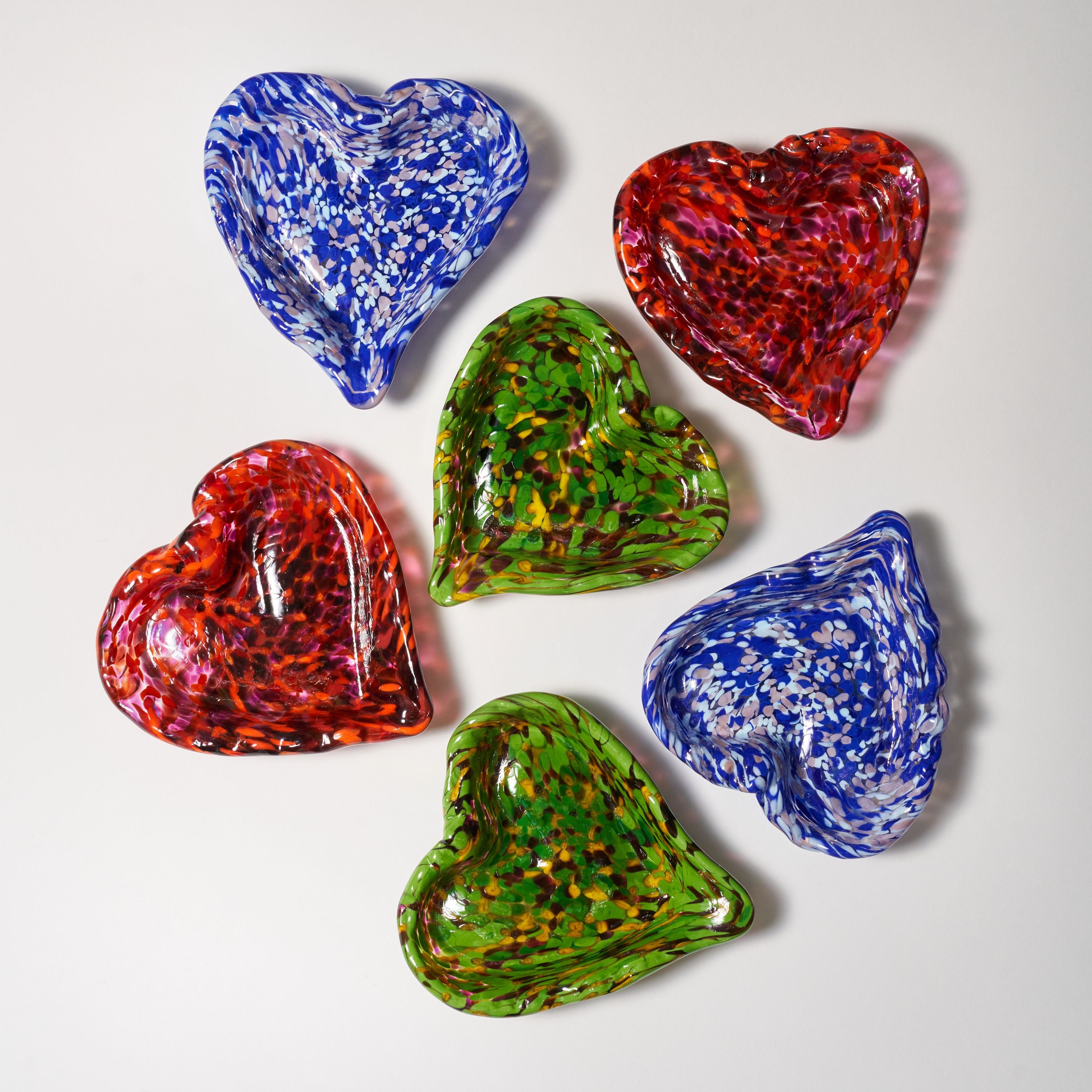 Six heart-shaped glass bowls in assorted vibrant colors, including blue, red, and green with marbled patterns, arranged on a light gray background.