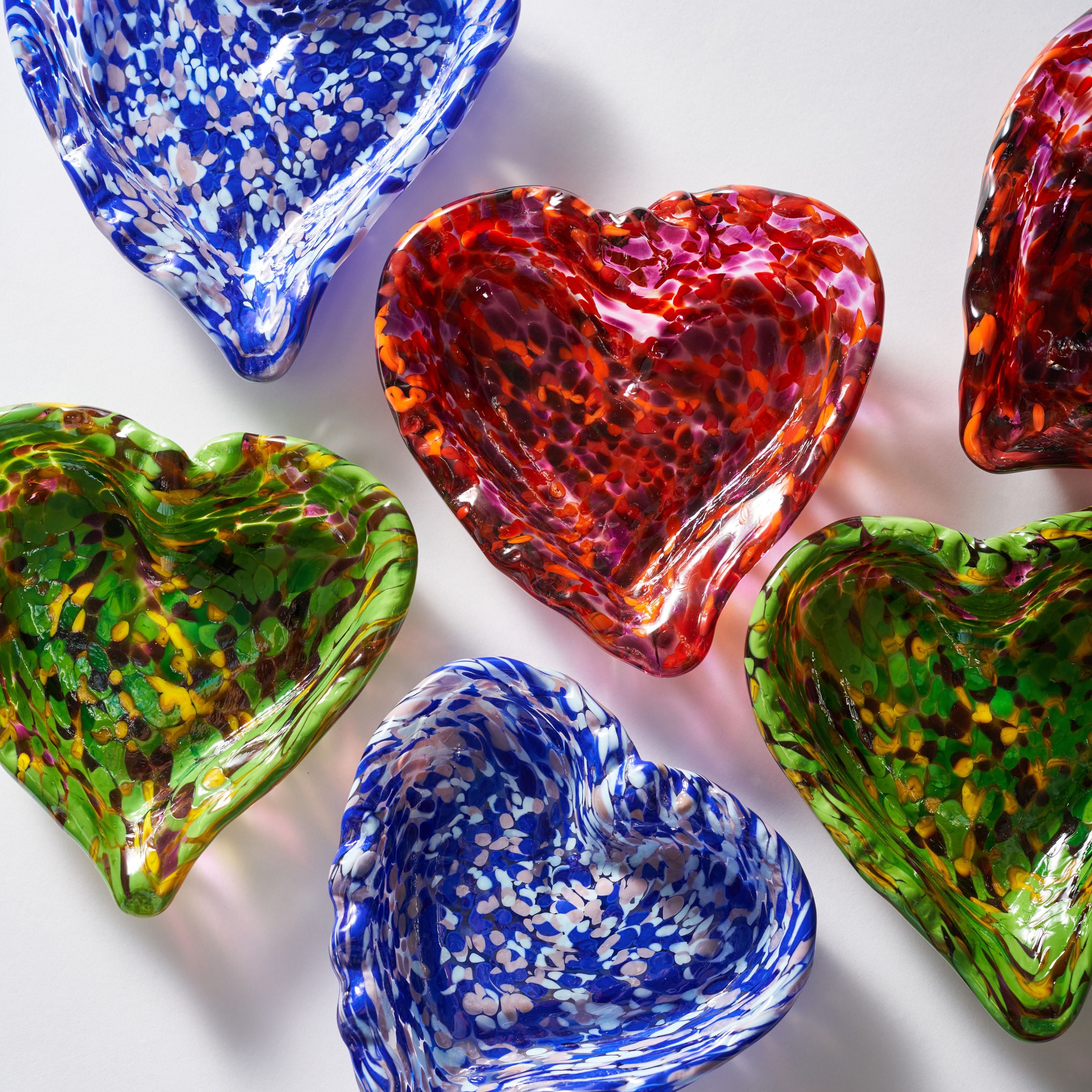 Six heart-shaped glass bowls in assorted colors and patterns, including blue-and-white speckled, red-and-orange marbled, and green-and-yellow mottled designs, arranged on a white background.
