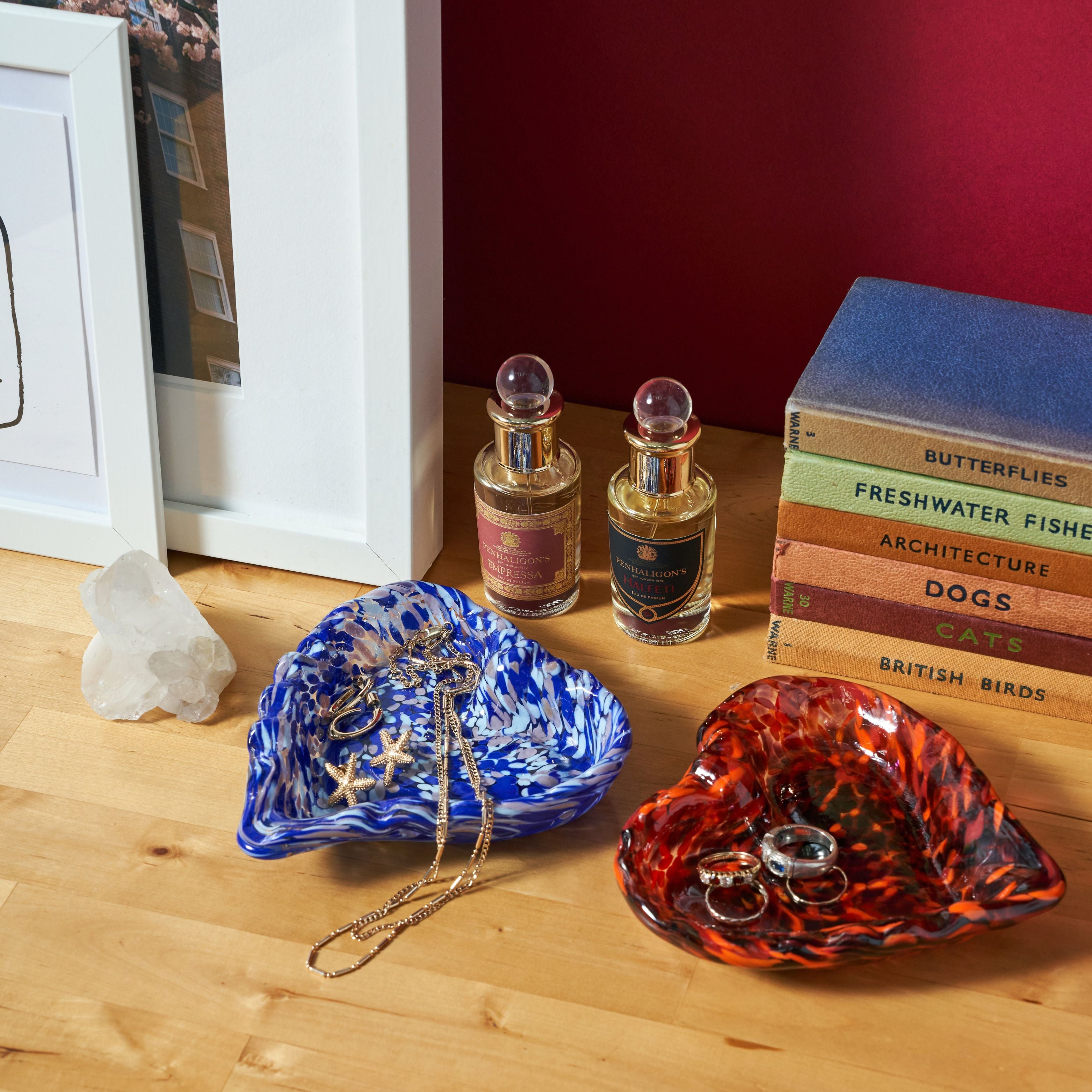 Two decorative glass jewelry trays on a wooden tabletop, one blue and white marbled and one orange and black marbled, styled with rings, necklaces, perfume bottles, books, and a framed print against a dark red wall.
