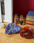 Two decorative glass jewelry trays on a wooden tabletop, one blue and white marbled and one orange and black marbled, styled with rings, necklaces, perfume bottles, books, and a framed print against a dark red wall.