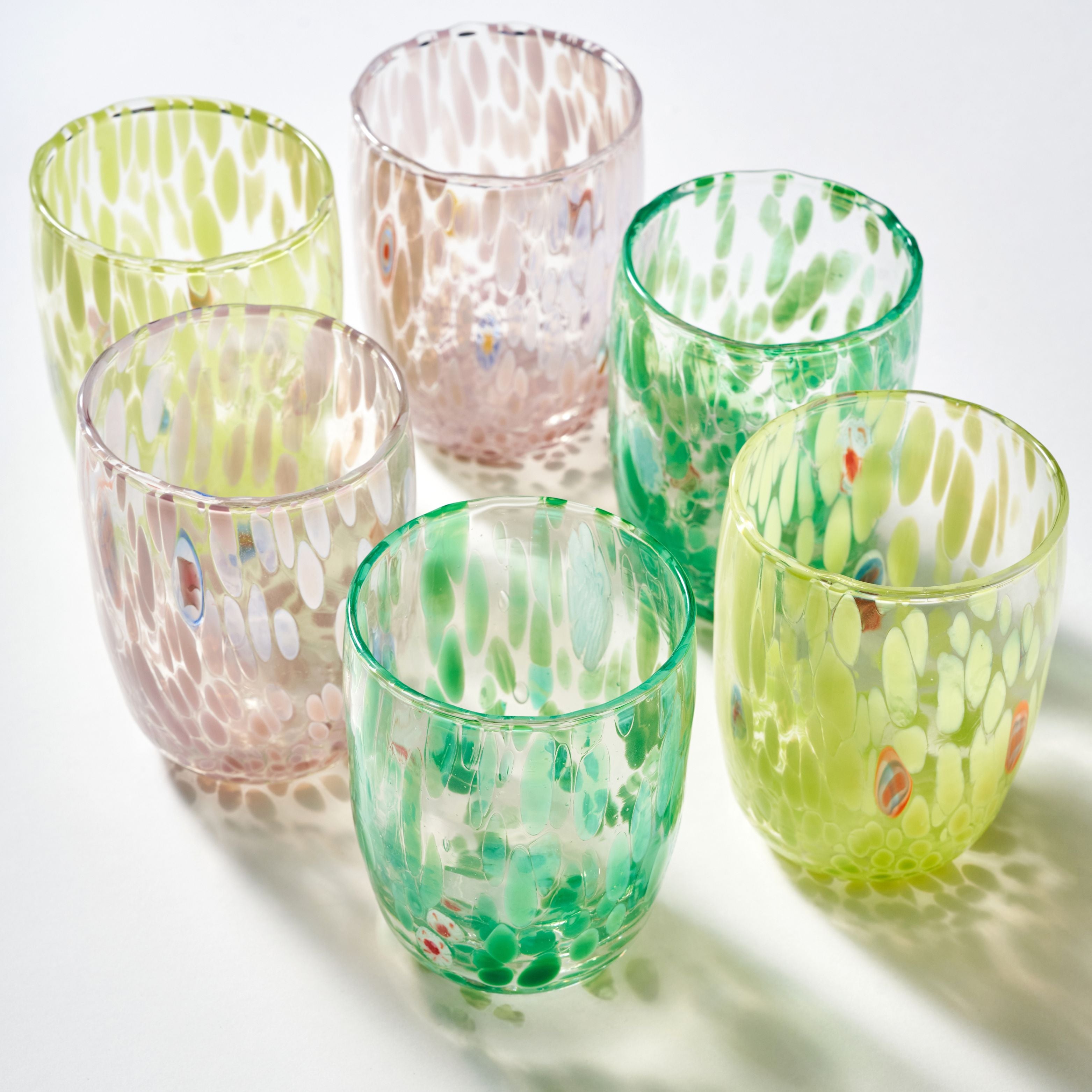 Six decorative drinking glasses with a textured, hammered glass finish in various pastel colors including green, pink, and yellow, arranged on a white surface with soft shadows.