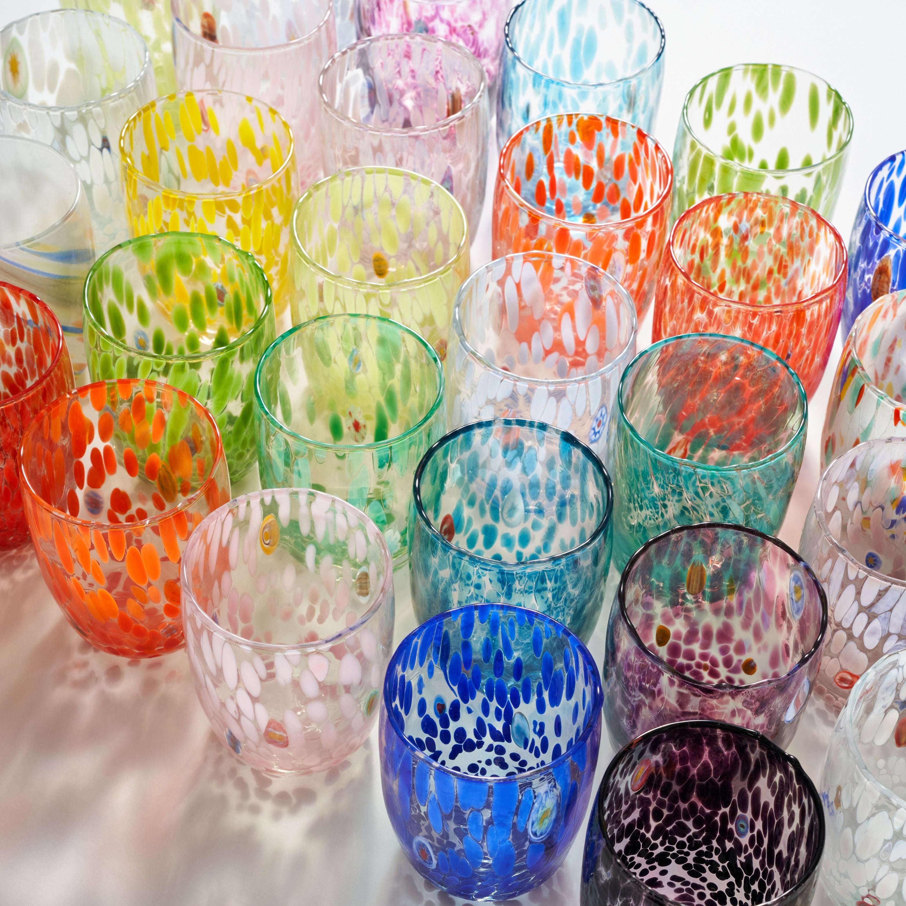 A collection of colorful hand-blown glass tumblers with white speckled patterns, arranged closely together on a light reflective surface.