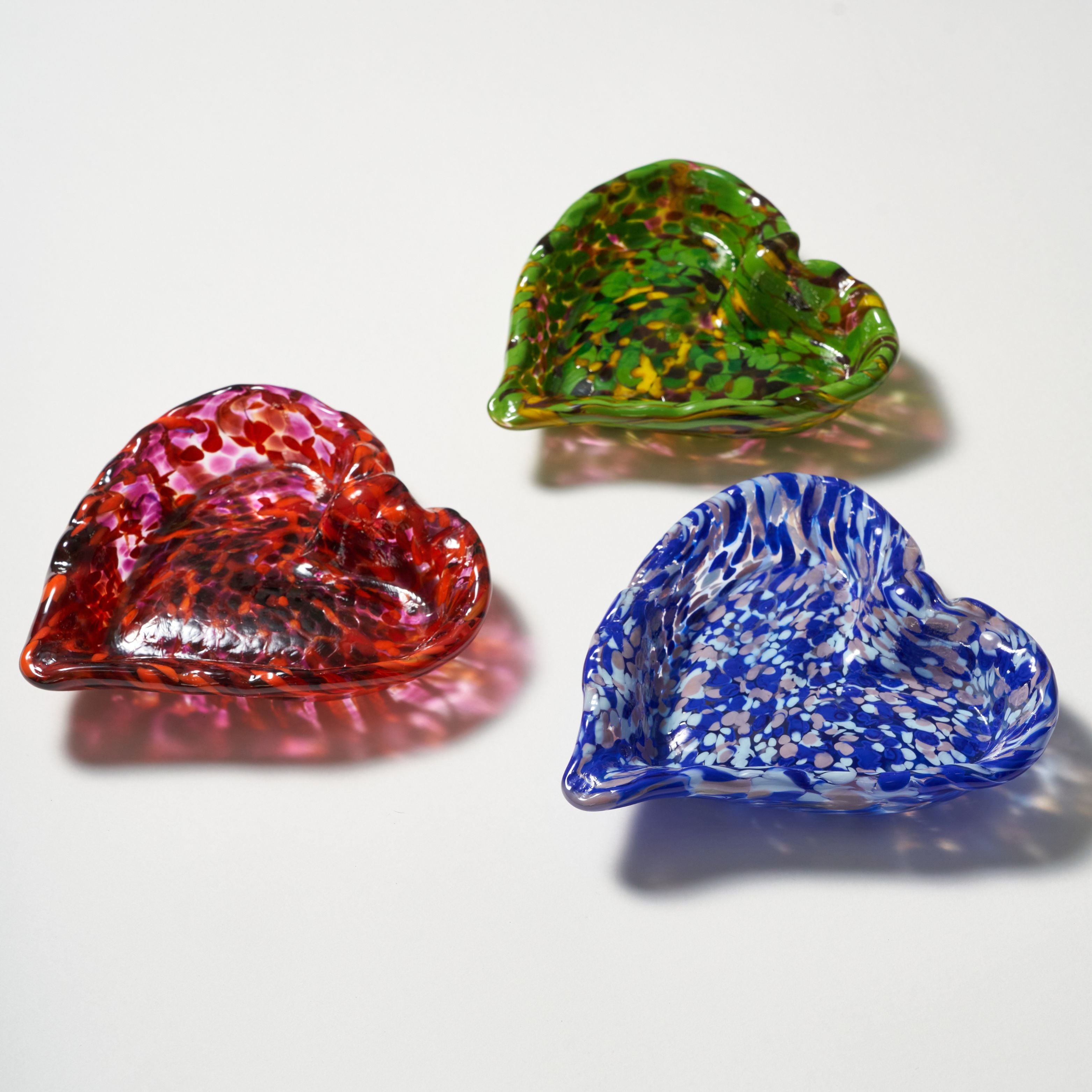 Three hand-blown glass bowls with a heart-like organic shape, shown in red, green, and blue colors with speckled patterns on a white background.